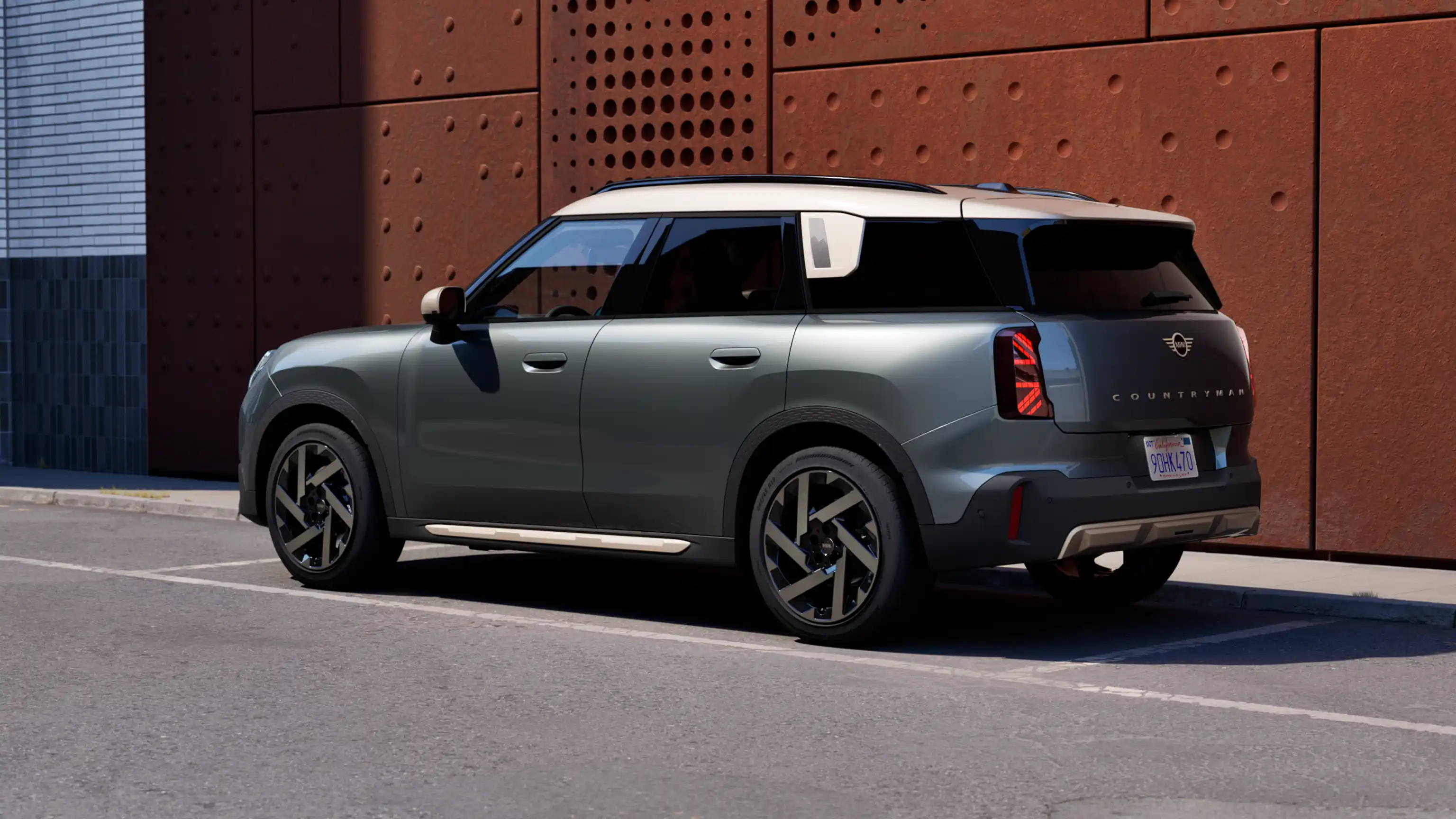 Rear three‑quarter view of a MINI Countryman in Sage Green Metallic parked beside a rust‑coloured wall.